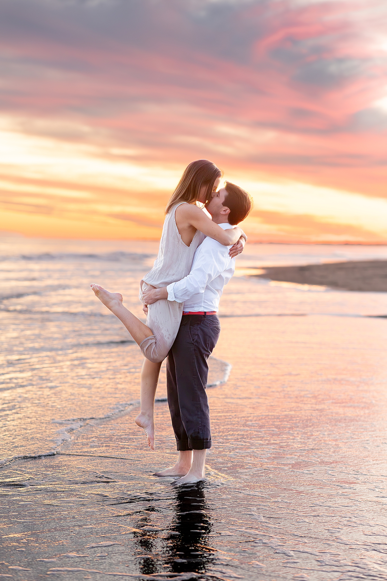 guy lifts his fiance as they kiss on the beach during sunset