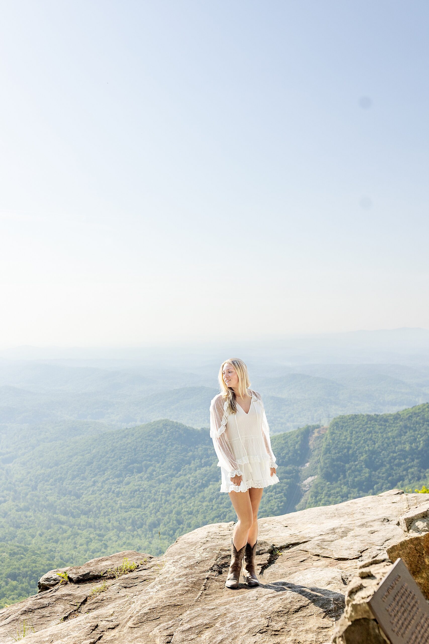 senior girl in white dress stands on rocky ledge with a beautiful mountain landscape behind her
