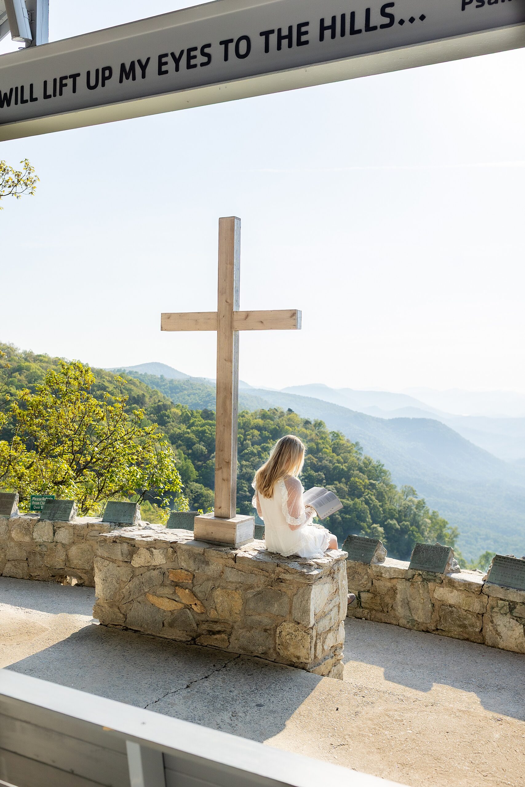 girl sits on stones by wooden cross and reads her bible