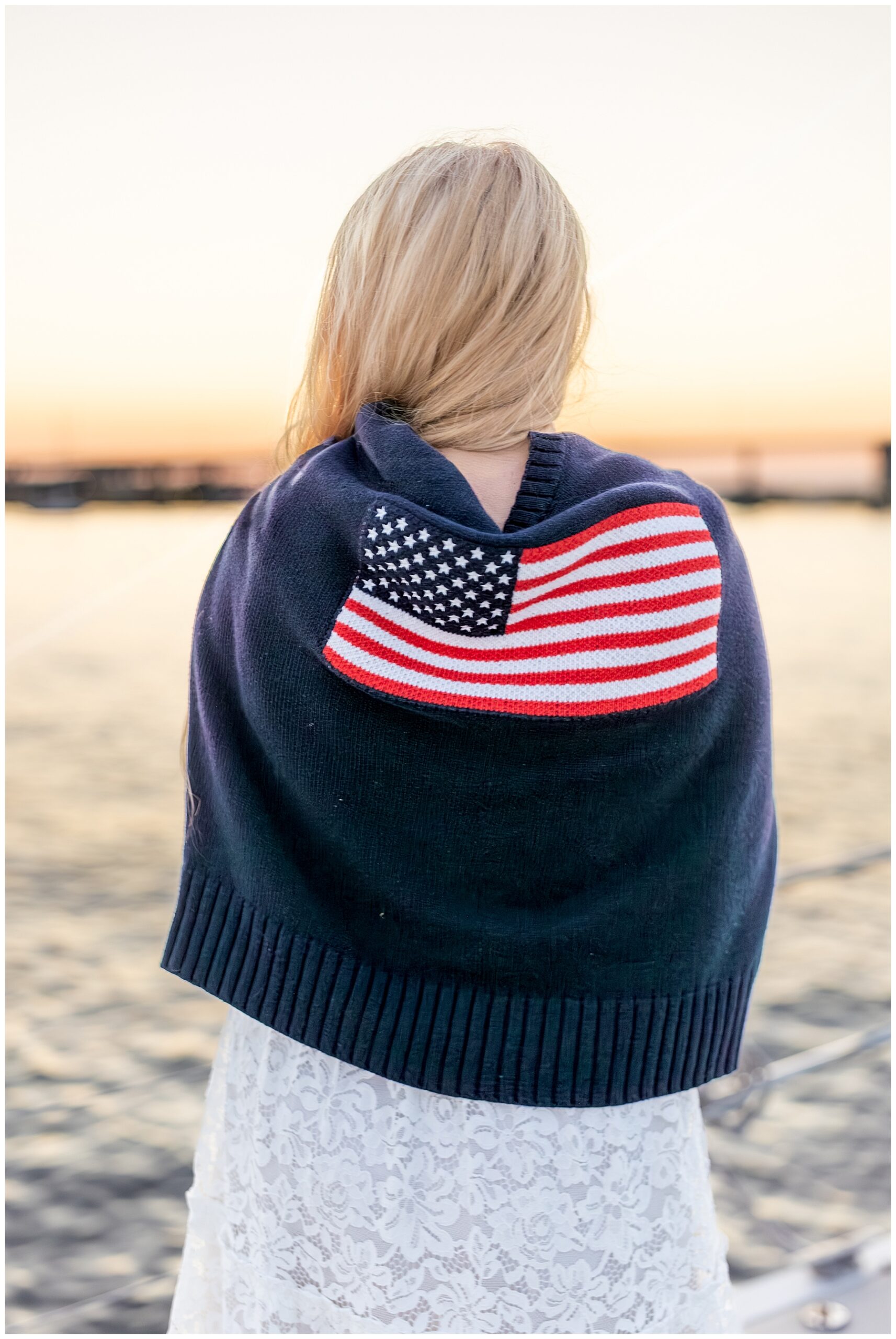 bride looks out at sunset while sailing on the water with American flag sweater around her arms 