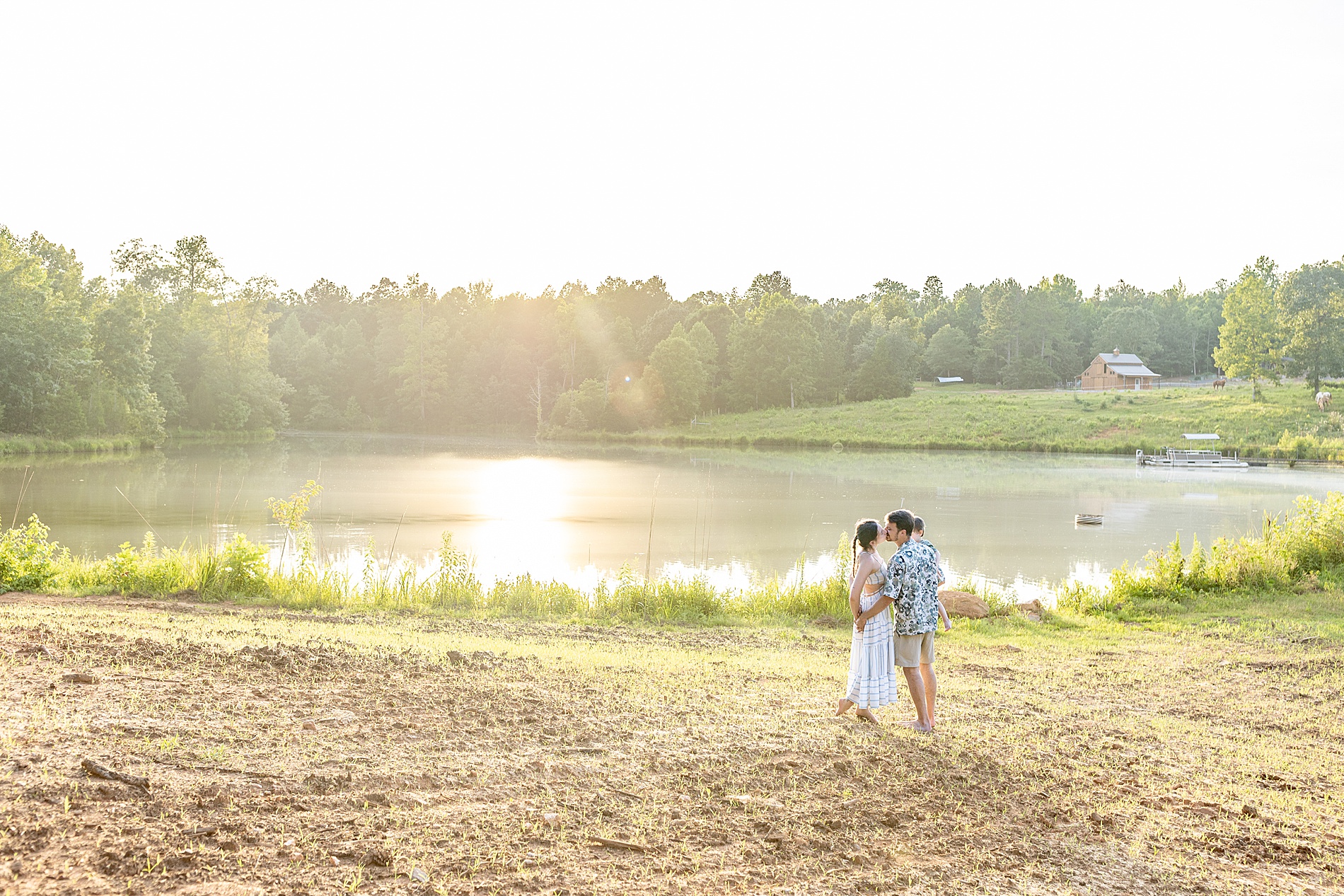 parents kiss on family farm by lake with sun shining overhead