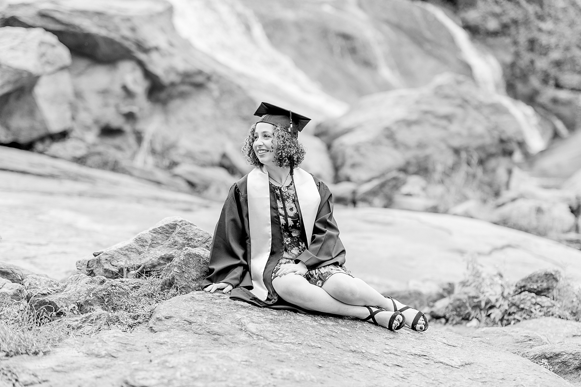 senior girl in cap and gown sits on rock by waterfall